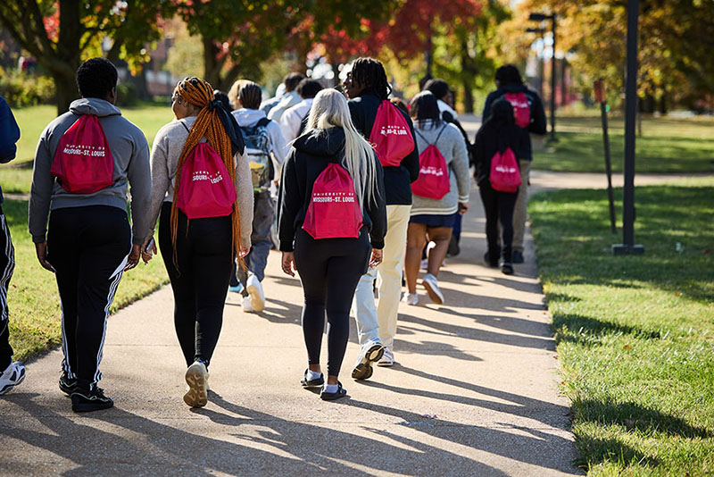 Group of students walking along a campus sidewalk with bright pink University of Missouri–St. Louis drawstring backpacks on a sunny autumn day.