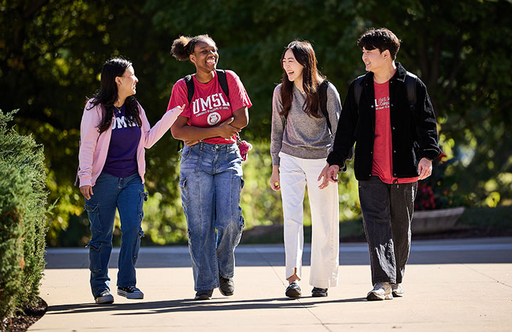 Four UMSL students walk together on a sunny campus path, chatting and smiling with backpacks on.