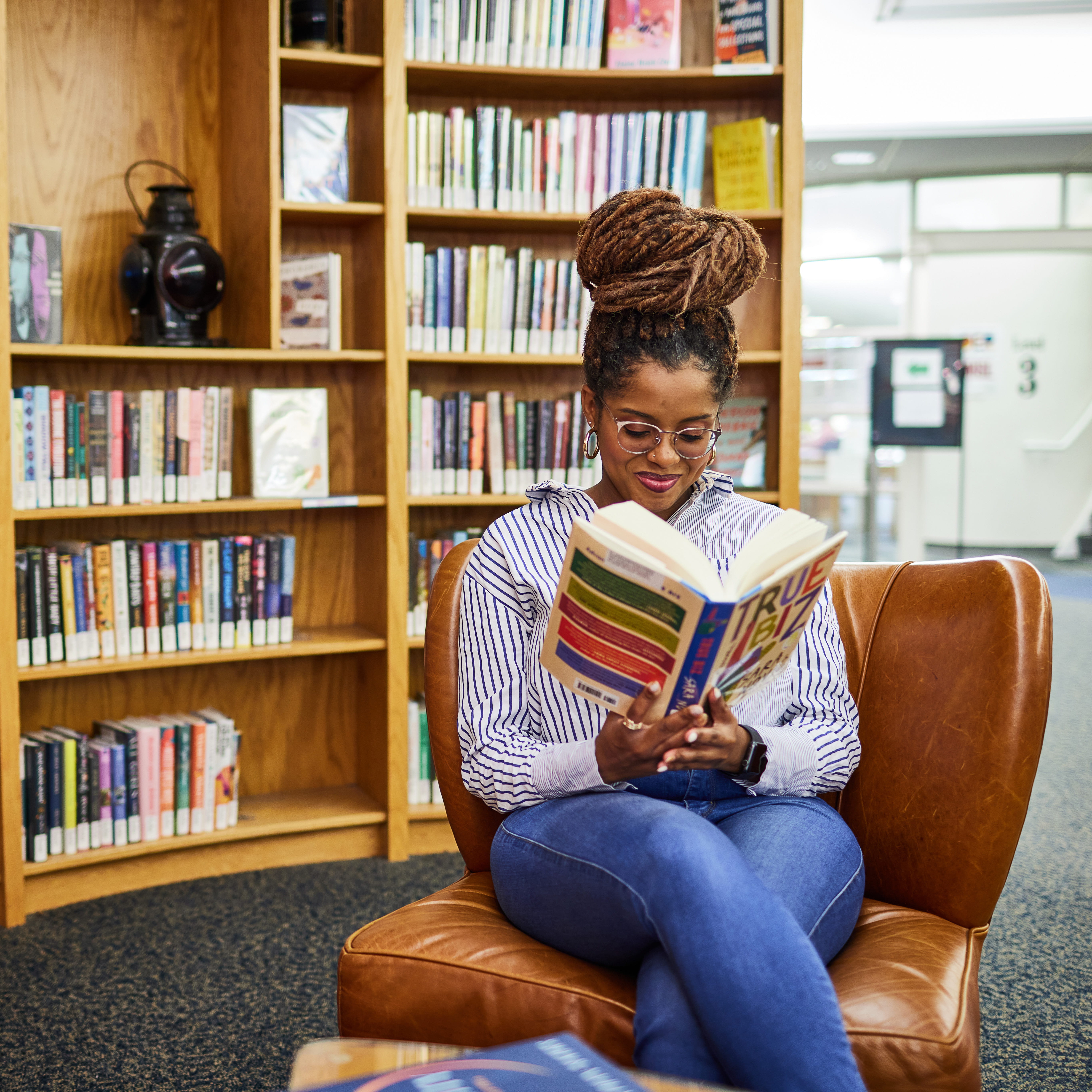 A woman with long, braided hair tied up in a bun sits comfortably in a leather chair at TJ Library, reading a colorful book titled True Biz. She wears glasses, a blue-and-white striped shirt and jeans, with bookshelves filled with novels in the background.