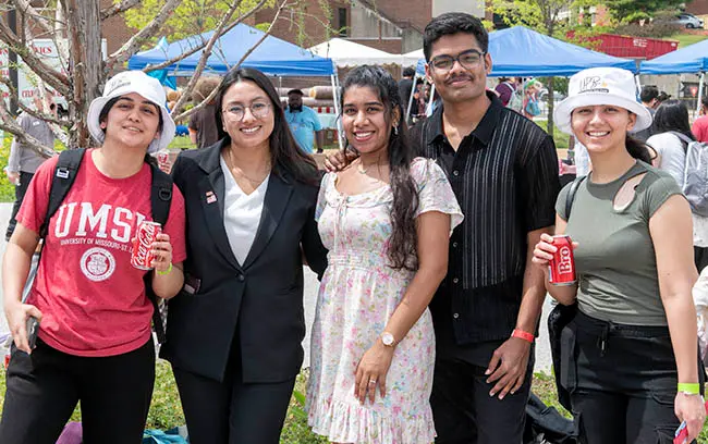 Group of five university students posing together outdoors at a campus event, smiling and holding drinks.