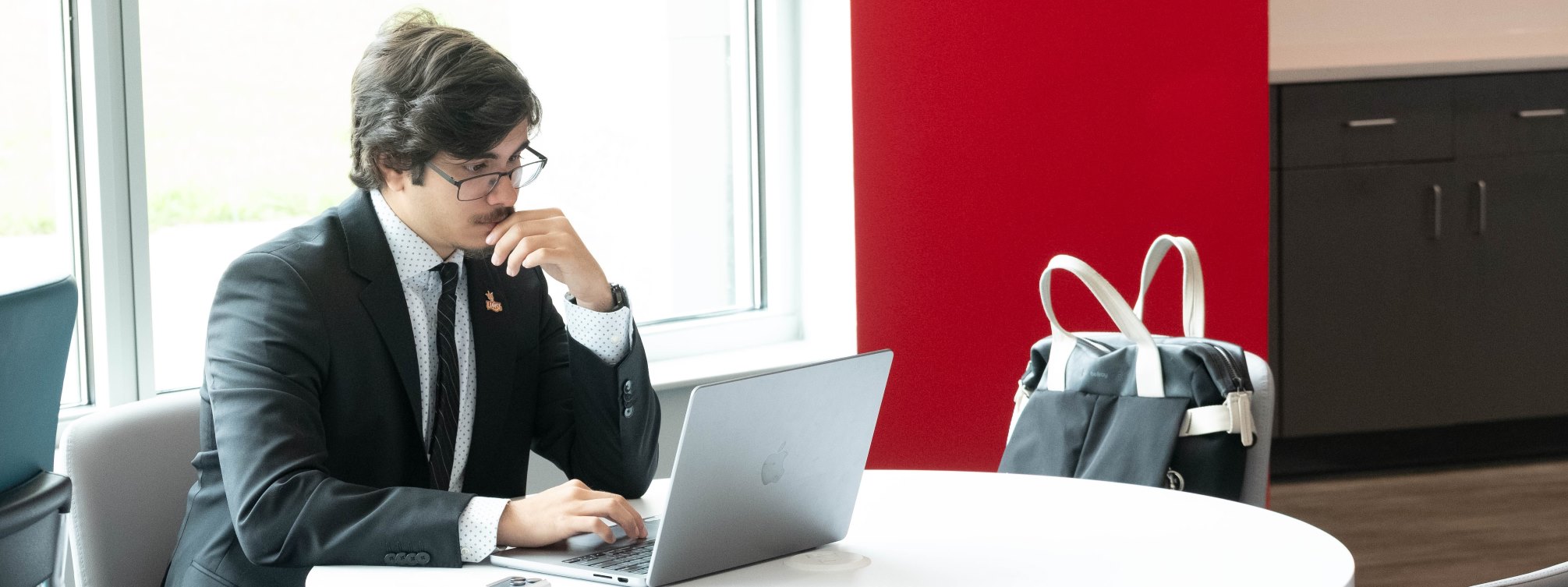 Student dressed in a suit looking at laptop with a backpack in a chair next to him