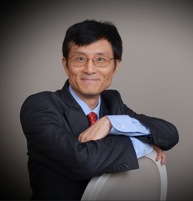 Smiling man in a dark suit and red tie, wearing glasses, leaning on the back of a chair against a neutral background.