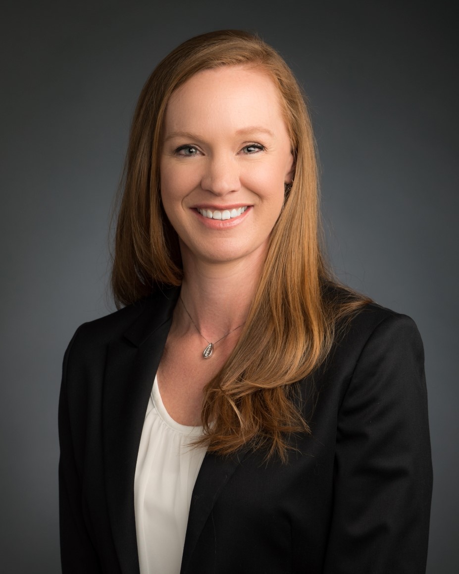 Professional headshot of a smiling woman with long red hair wearing a black blazer and white blouse against a gray background.
