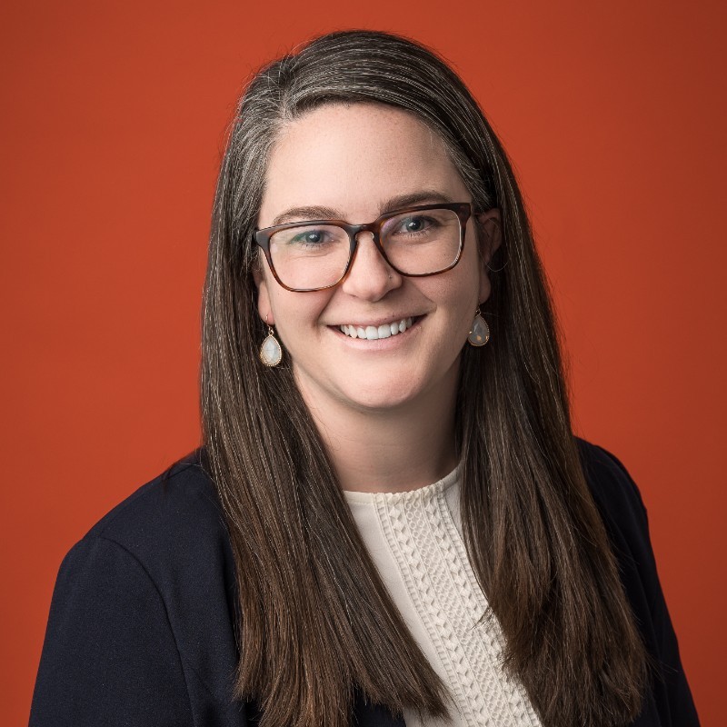 Smiling woman with long hair and glasses in a professional headshot against an orange background.