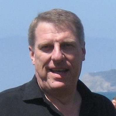 Close-up portrait of a man outdoors with a blue sky and distant mountains in the background.