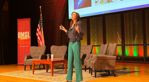 a women talking on a stage with a banner on her right side and chairs and a table behind her