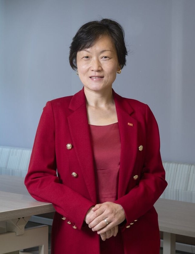 Portrait of Shu Schiller in a red blazer standing indoors with hands clasped.