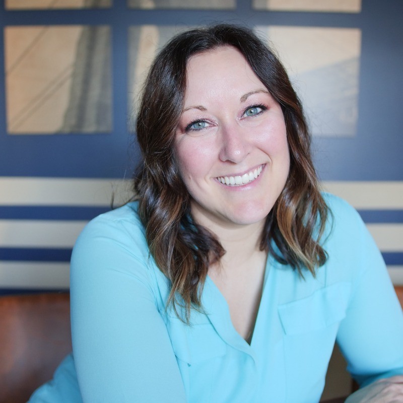 Smiling woman with shoulder-length wavy hair wearing a light blue top, seated indoors in front of a windowed wall.