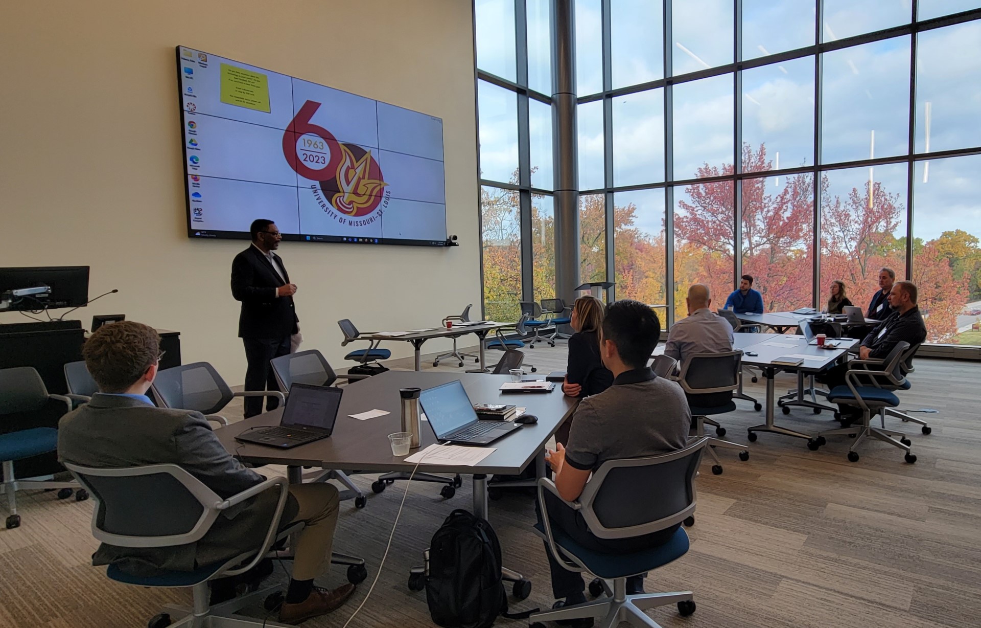 A presenter leads a meeting in a modern conference room with large windows showcasing an autumn landscape.