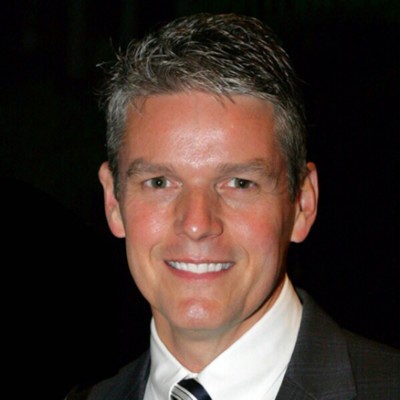Headshot of a Joe Stoff, smiling man in a suit and tie against a dark background.