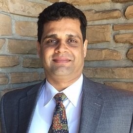 Headshot of Vish Tripathi in a suit with a patterned tie, smiling against a textured stone wall.