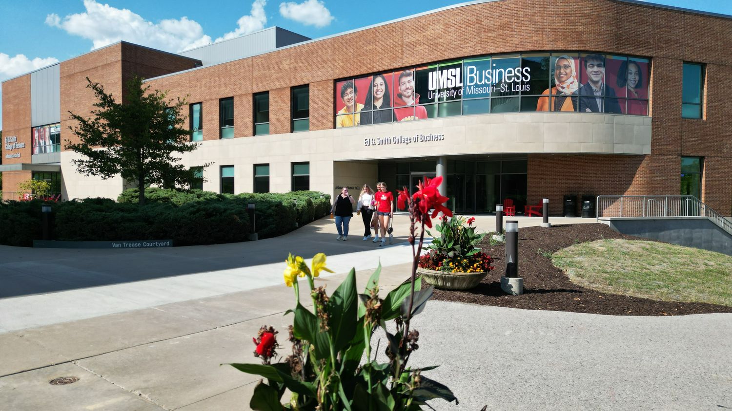 a campus building with windows and students walking out of it with a flower in the front of the photo