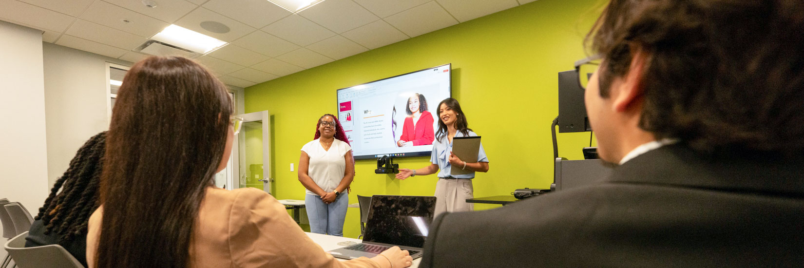 Two presenters at the front of a modern classroom deliver a presentation while participants sit at tables with laptops, facing a large wall-mounted screen displaying slides against a bright green wall