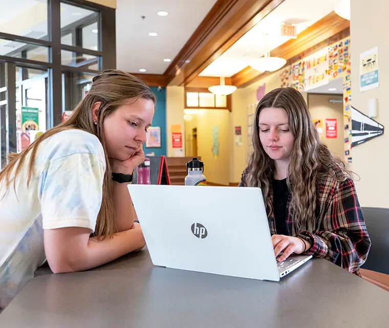 Two students collaborating in a study area, focused on a laptop open on a table.