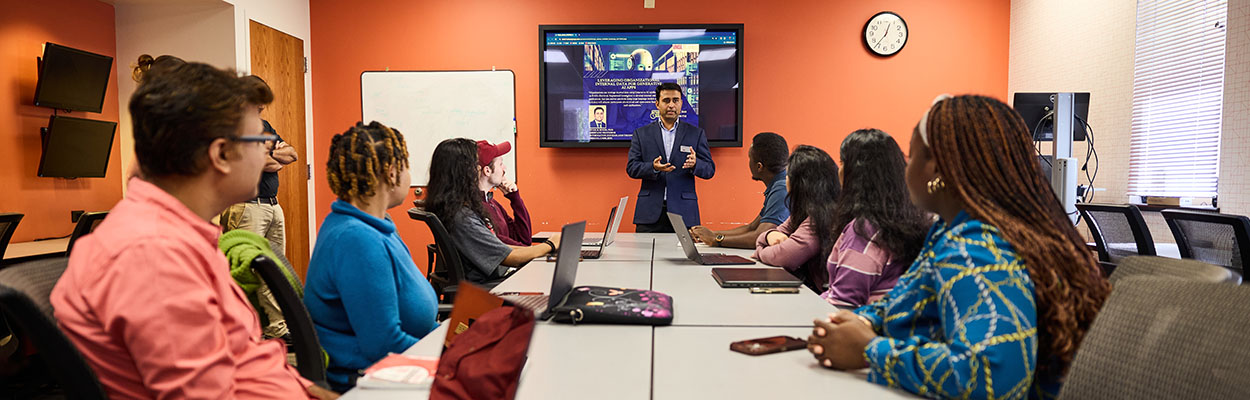 professor standing by a tv display talking to students seated around a table
