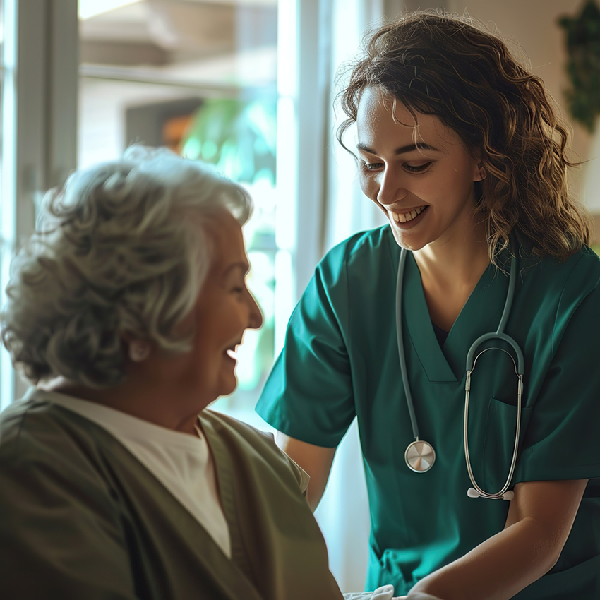 Nurse in scrubs smiling while assisting an elderly woman in a bright care facility.