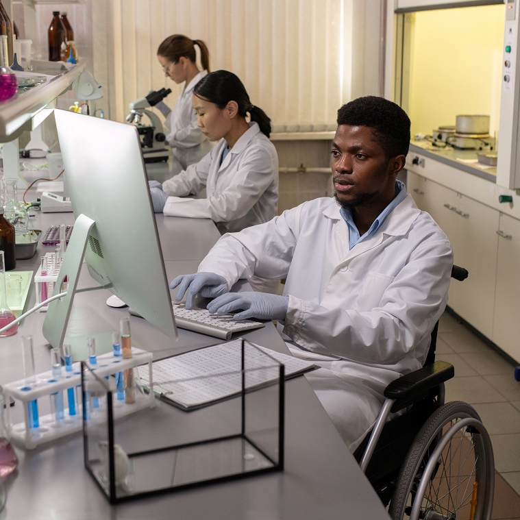 A scientist in a wheelchair working at a computer in a laboratory, with two colleagues conducting research in the background.