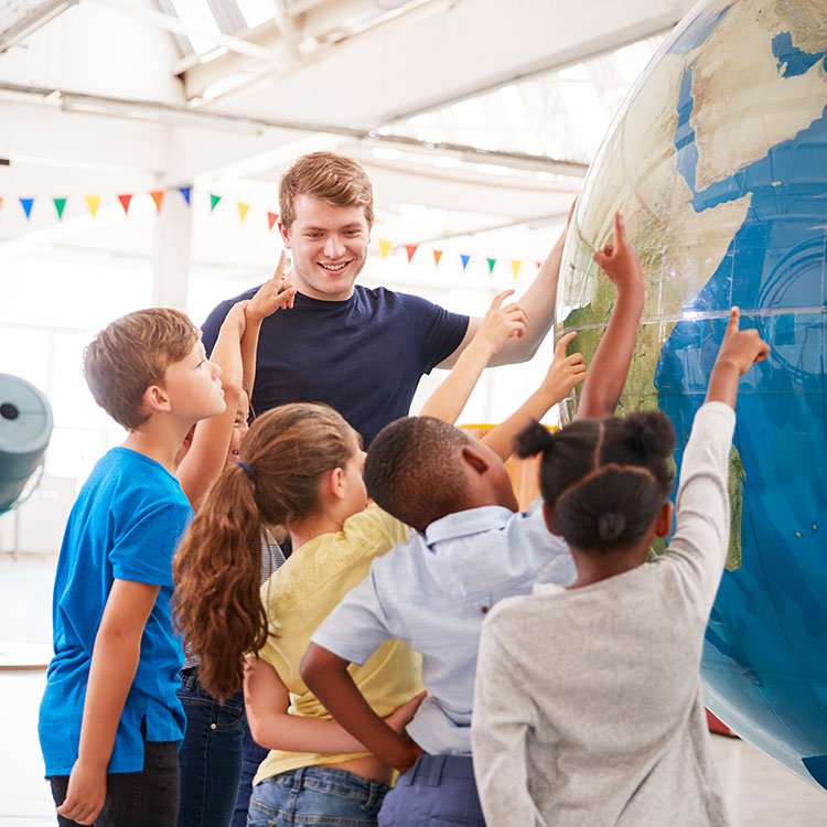 A teacher engages a group of diverse children as they excitedly point to locations on a large globe.