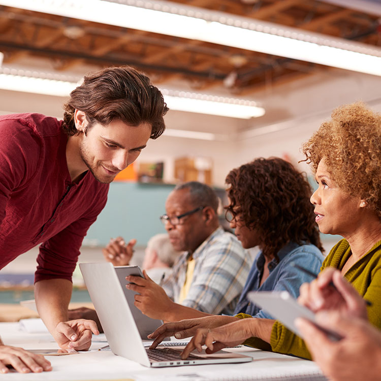 A diverse group of people engaged in a collaborative work session, with one person presenting information on a laptop while others are discussing and using various devices.