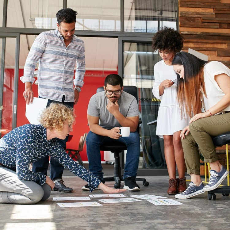 A group of five diverse professionals collaborates around a floor layout, discussing ideas and reviewing documents in a modern office setting.