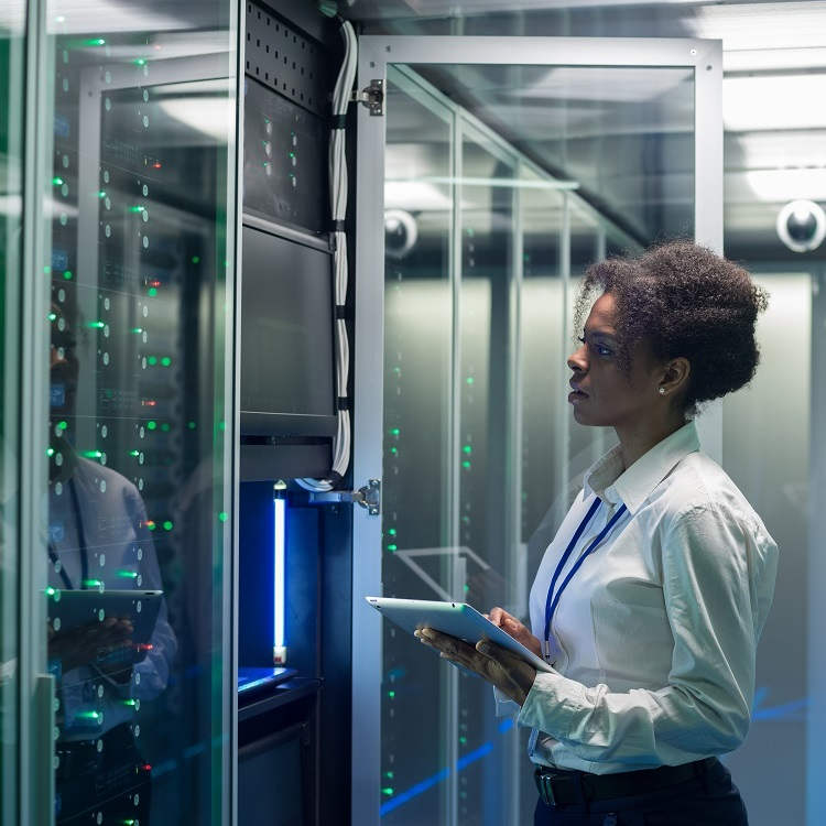 A technician in a server room monitors equipment while holding a tablet.