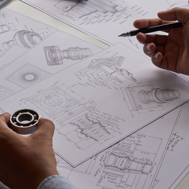 Technical drawings and schematics of machine components are displayed on a table, with hands holding a bearing and a pen.