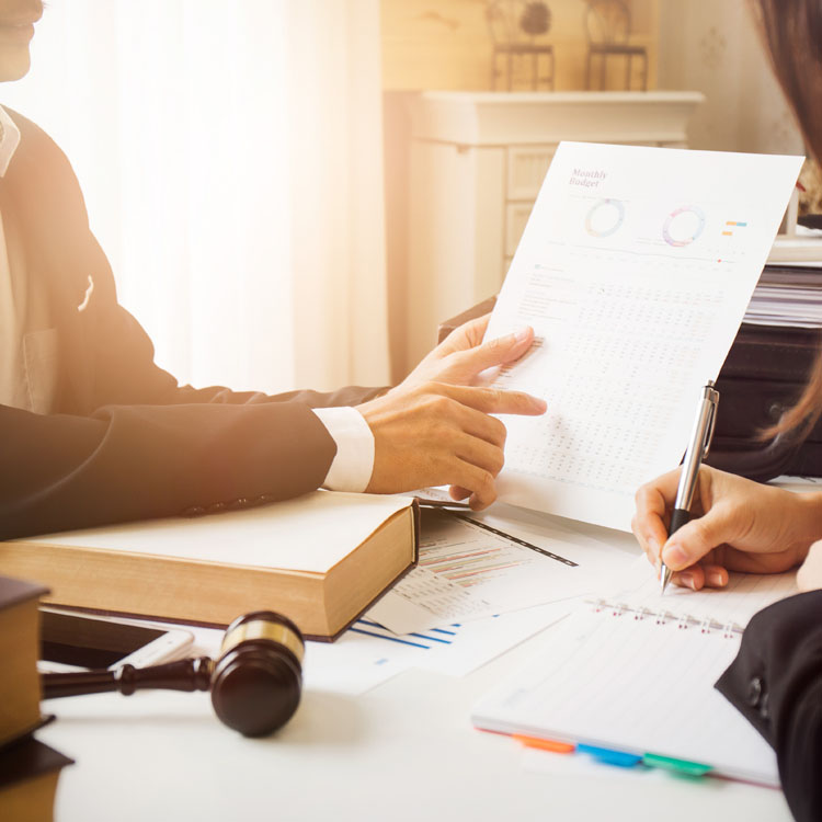 A business meeting in progress, featuring a person pointing at a financial report while another takes notes, with legal books and a gavel visible on the table.