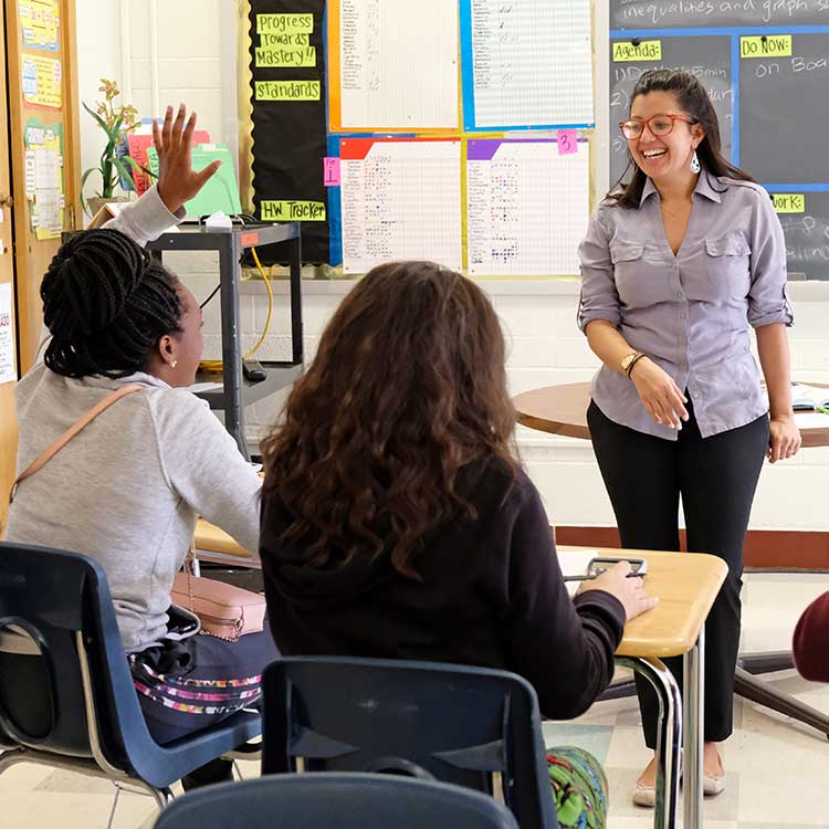 A teacher actively engaging with students in a classroom setting, where one student has raised their hand to participate.