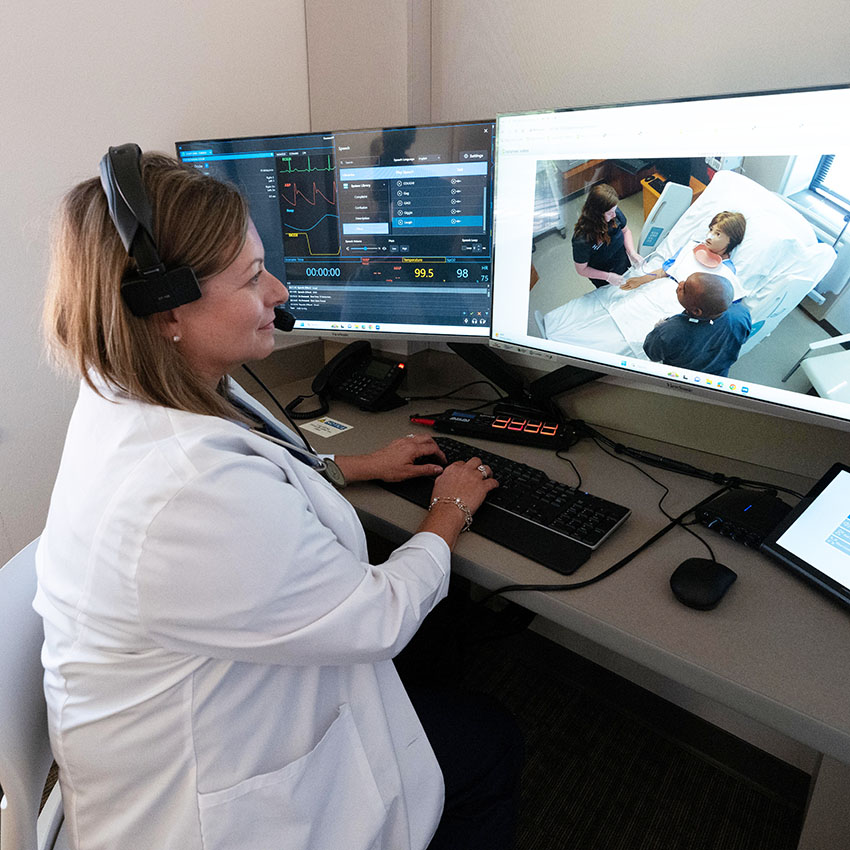 Nursing Instructor using a headset while monitoring patient care on dual screens in a clinical setting.