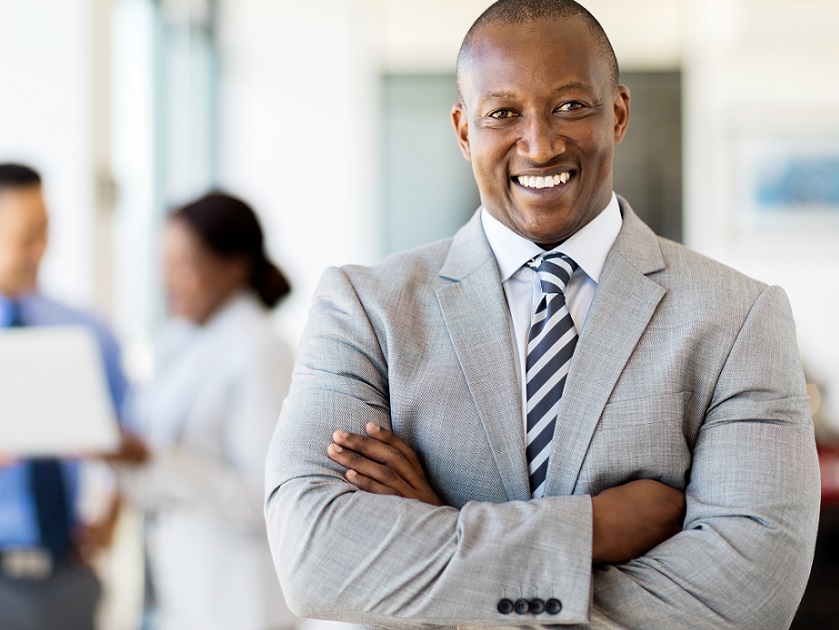 Confident businessman wearing a gray suit and striped tie, standing with arms crossed in a professional setting.