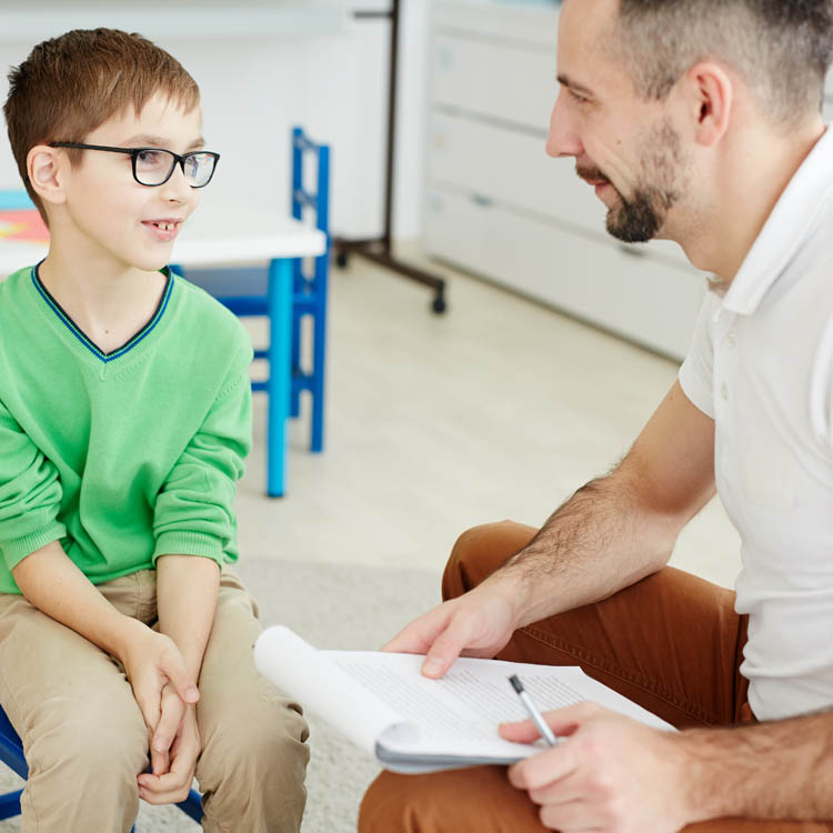 A child with glasses engages in a conversation with an adult, likely a teacher or counselor, in a classroom setting.