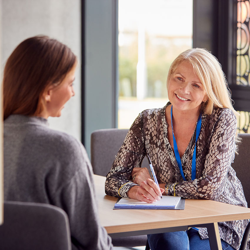 A woman with blonde hair smiles while taking notes during a conversation with another woman in a casual meeting setting.