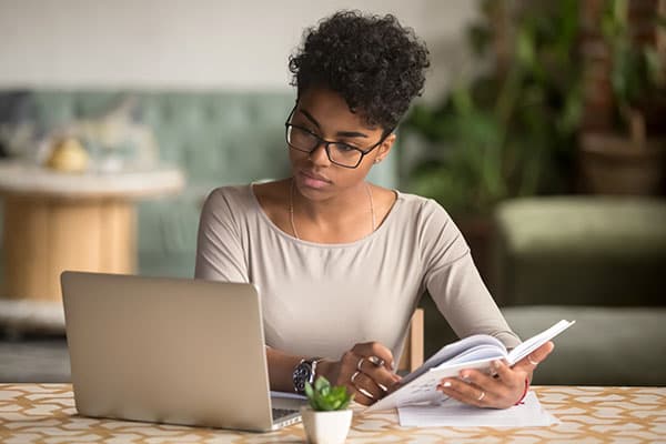 A woman with curly hair wearing glasses engages in her studies while working on a laptop, surrounded by indoor plants.