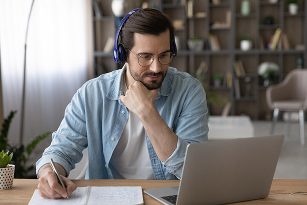 A man wearing headphones is focused on his laptop while taking notes