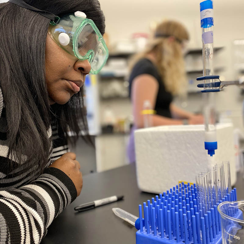 A scientist in safety goggles closely observes a lab experiment involving pipettes and test tubes.