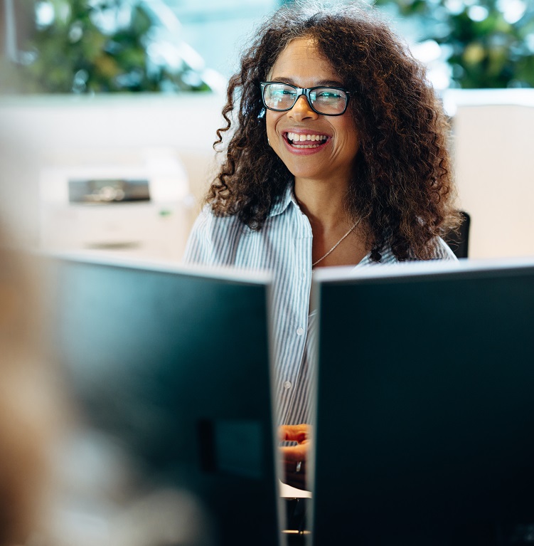 A smiling woman with curly hair and glasses sitting at a desk, engaged with a computer, suggesting a positive work environment.