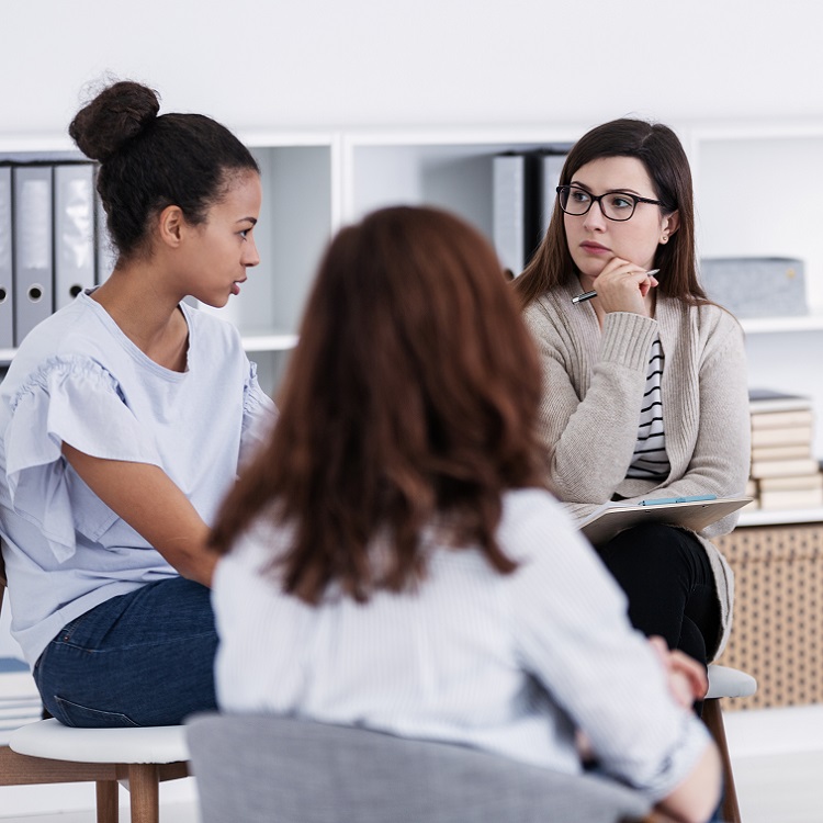 A group of three women engaged in a thoughtful discussion in a modern meeting space.
