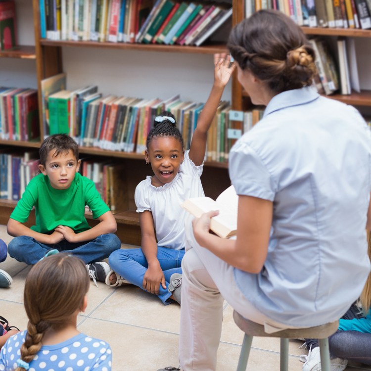 A teacher reads to a group of engaged children in a library, with one girl raising her hand to answer a question.