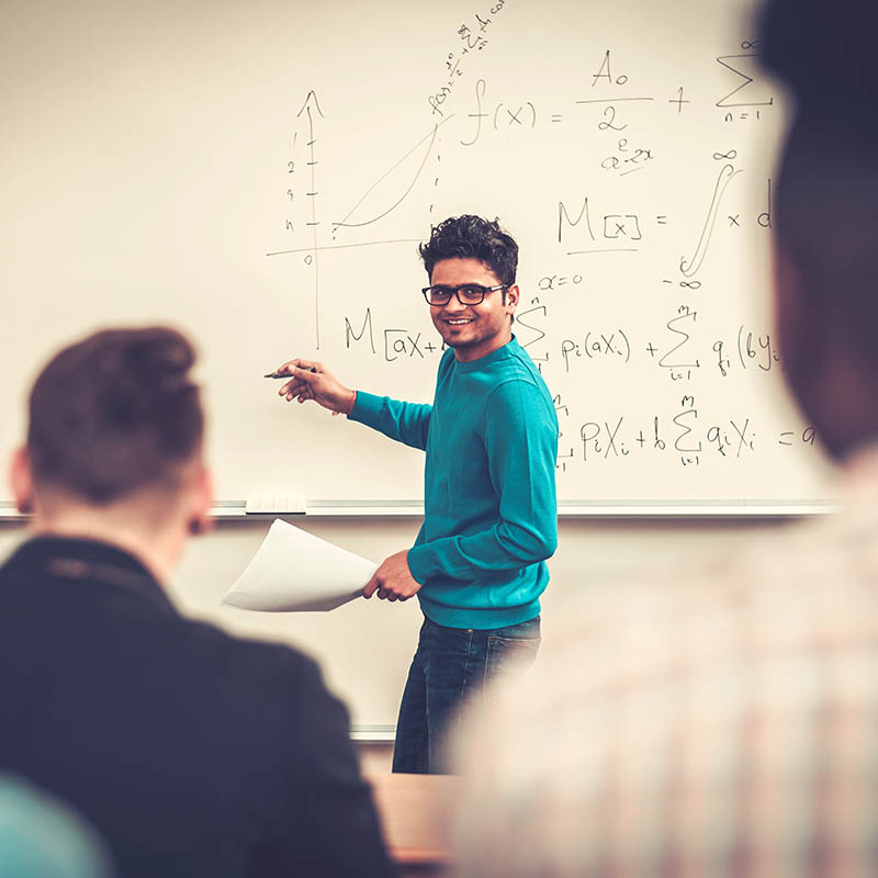 A young teacher in a blue sweater explains mathematical concepts on a whiteboard to a classroom of students.