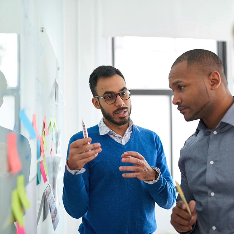 Two professionals engaged in a discussion while reviewing notes and ideas on a glass wall.