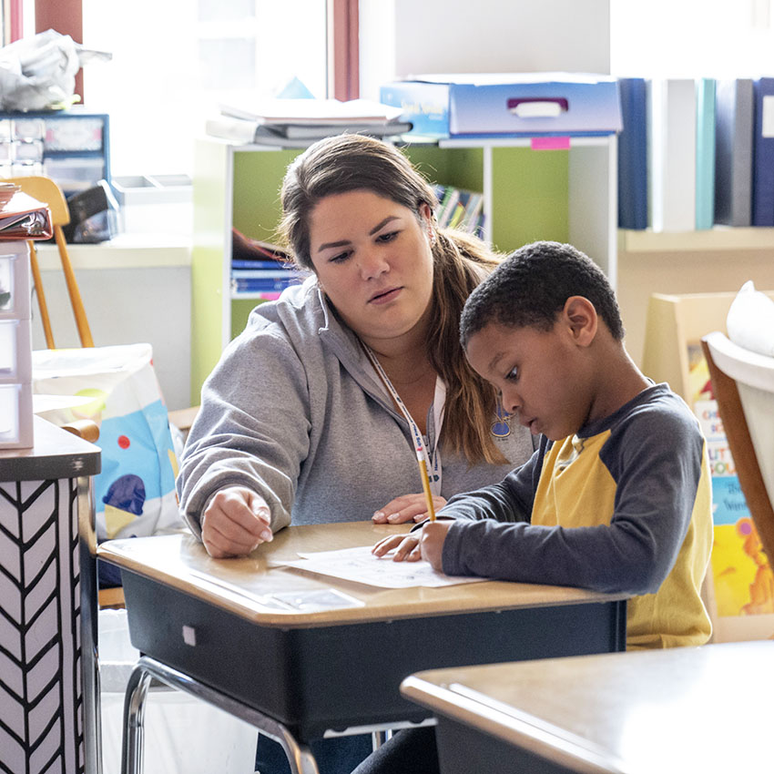 A teacher assists a young student with their work at a classroom desk.