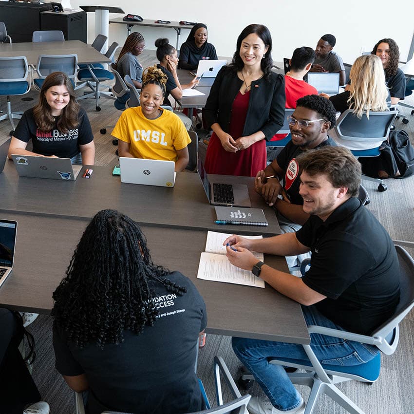 A group of UMSL students collaborates around a table in a classroom setting, with a faculty member engaging with them.