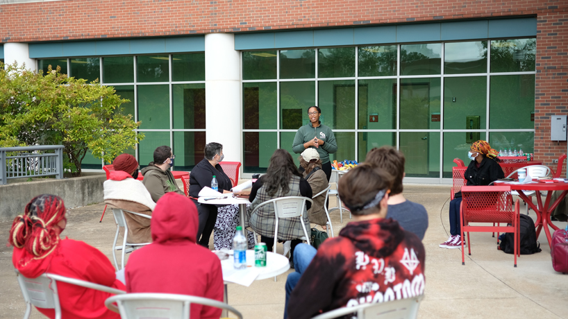 Groups of people sit at tables outdoors and watch a person speak