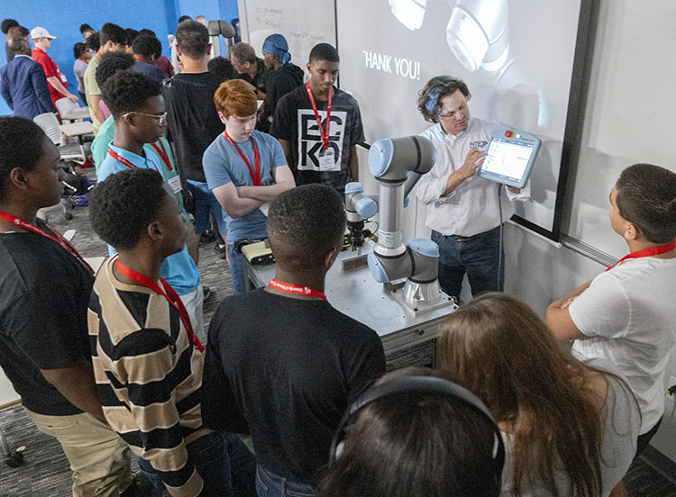 A group of students attentively engages with an instructor demonstrating robotics technology using a touchscreen interface.
