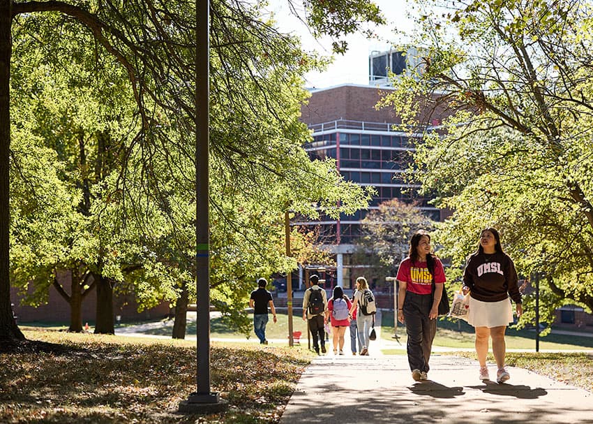 UMSL students walking on a campus path surrounded by trees, with the science complex in the background.