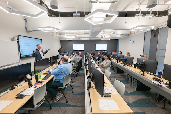 Instructor presenting to a group in a modern computer lab classroom with rows of desktop monitors and students seated at workstations.