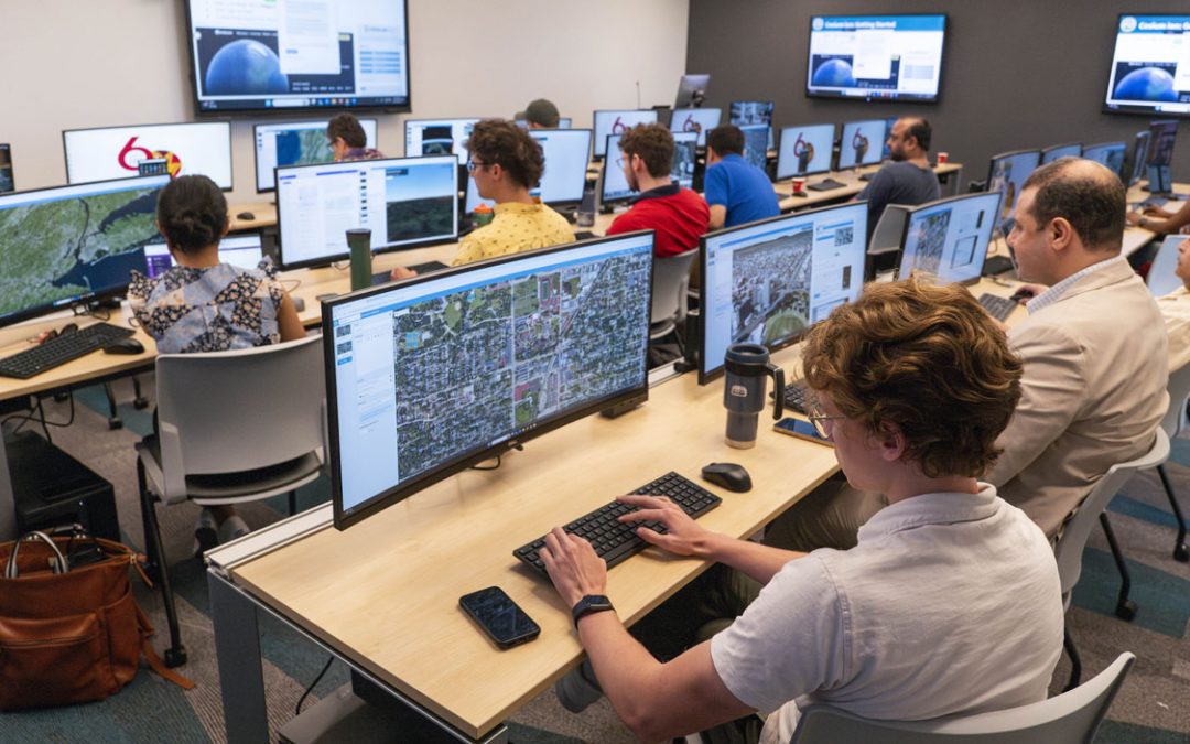 Students work together at desktop computers in a classroom lab, with one student smiling while talking to a classmate.