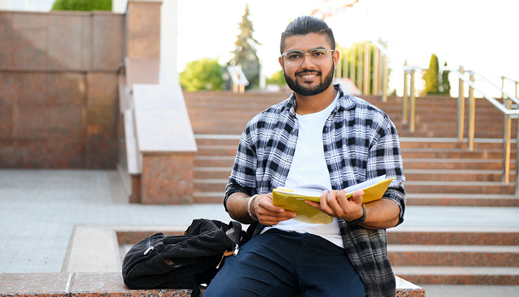 student sitting outside on a low wall with a book