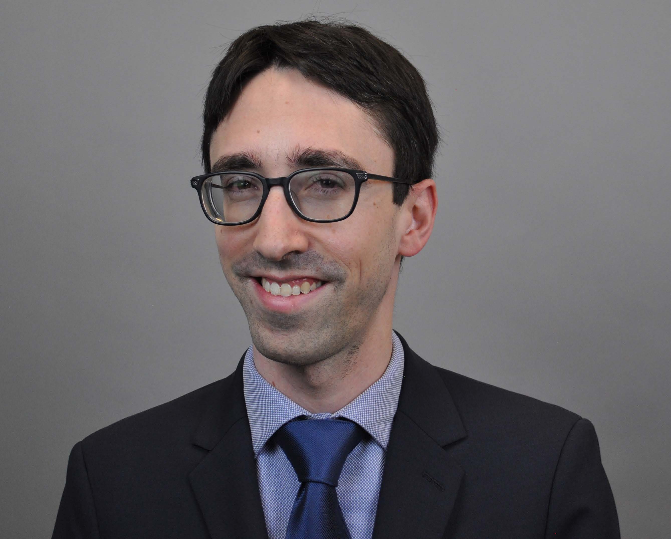Head-and-shoulders portrait of a smiling man with dark hair and black-framed glasses, wearing a suit and blue tie against a plain gray background.
