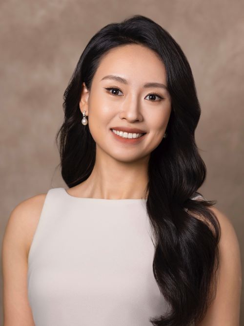 Studio headshot of a smiling woman with long dark hair, wearing pearl earrings and a light sleeveless top against a neutral background.